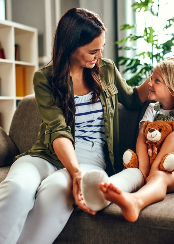 Ein kleines Mädchen mit eingegipstem Bein und Teddybär auf dem Schoß wird von ihrer Mutter auf dem Sofa getröstet.