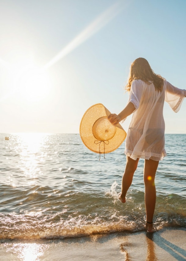 Eine junge Frau im weißen Sommerkleid und mit Strohhut in der Hand planscht mit ihrem Fuß im Wasser an einem Sandstrand am Meer bei Sonnenuntergang